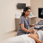 A professional female ultrasound technician in grey scrubs performing an abdominal scan on a female patient lying on an exam table in a medical clinic.